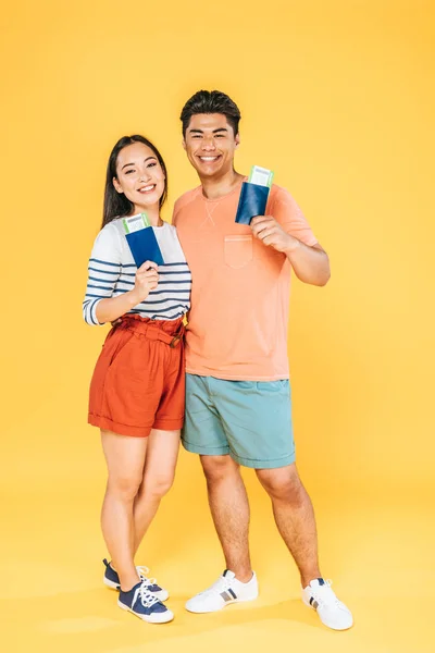 Due turisti asiatici felici in possesso di passaporti e biglietti aerei mentre sorridono alla fotocamera su sfondo giallo — Foto stock
