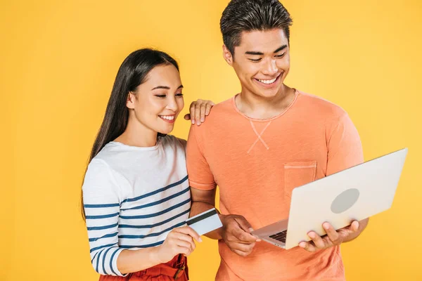 Jovem asiático homem usando laptop perto namorada segurando cartão de crédito isolado no amarelo — Fotografia de Stock