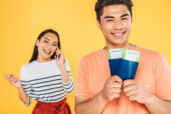 Selektiver Fokus eines glücklichen asiatischen Mannes mit Pässen und Flugtickets in der Nähe einer fröhlichen Freundin, die auf einem Smartphone isoliert auf gelb spricht — Stockfoto