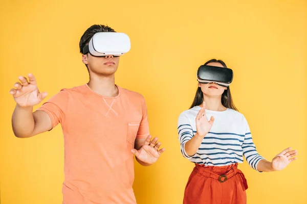 Joven hombre y mujer haciendo gestos mientras usa auriculares de realidad virtual sobre fondo amarillo - foto de stock