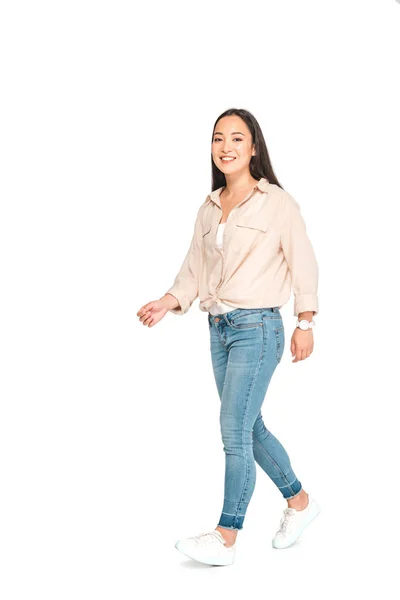 Atractiva mujer asiática en pantalones vaqueros azules mirando a la cámara mientras camina sobre fondo blanco - foto de stock