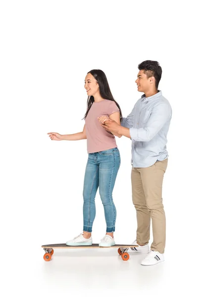 Smiling asian man helping girlfriend riding longboard on white background — Stock Photo