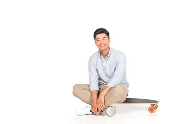 Sonriente asiático hombre sentado en longboard y mirando la cámara en blanco fondo - foto de stock