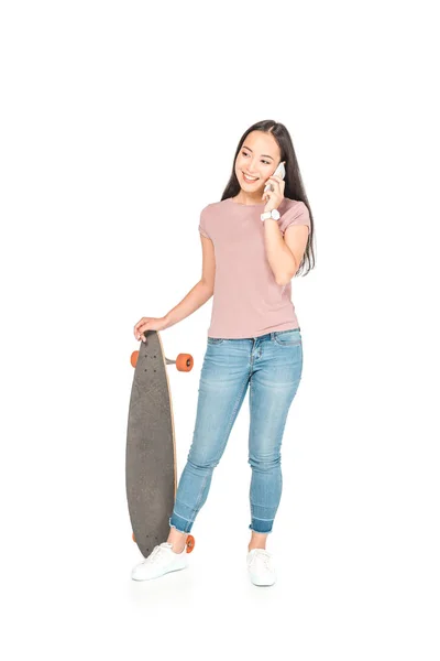 Pretty asian girl talking on smartphone while holding longboard on white background — Stock Photo