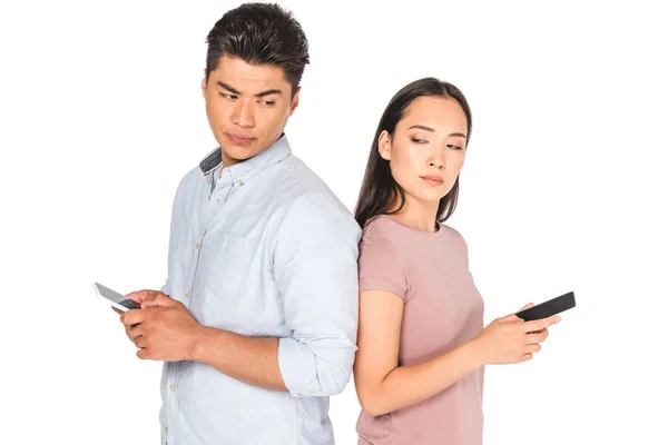 Displeased asian couple looking at each other while using smartphones isolated on white — Stock Photo