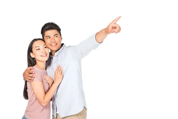 Happy asian man hugging smiling girlfriend while pointing with finger isolated on white — Stock Photo