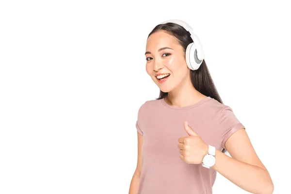 Felice donna asiatica ascoltando musica in cuffia, mostrando pollice in su e sorridendo alla fotocamera isolata su bianco — Foto stock