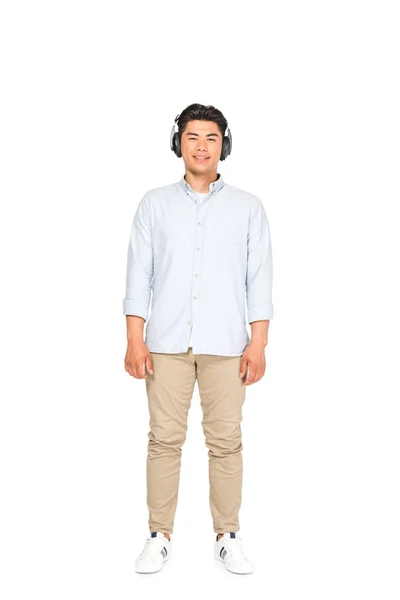 Longitud completa vista de guapo asiático hombre escuchando música en auriculares y sonriendo a la cámara en blanco fondo - foto de stock