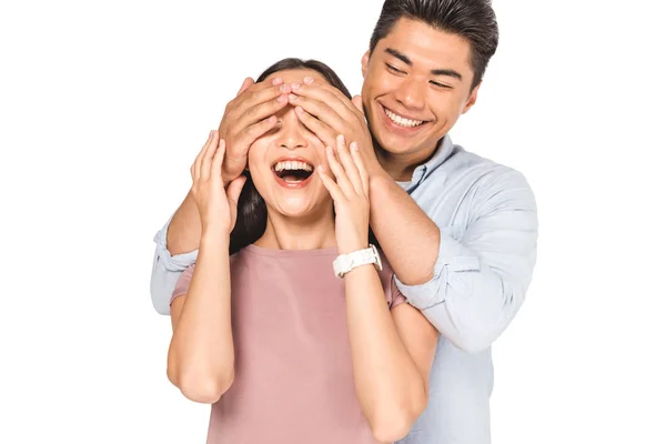 Cheerful asian man closing eyes of smiling girlfriend with hands isolated on white — Stock Photo