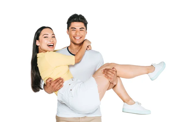 Cheerful asian man holding happy girlfriend on hands and smiling at camera isolated on white — Stock Photo