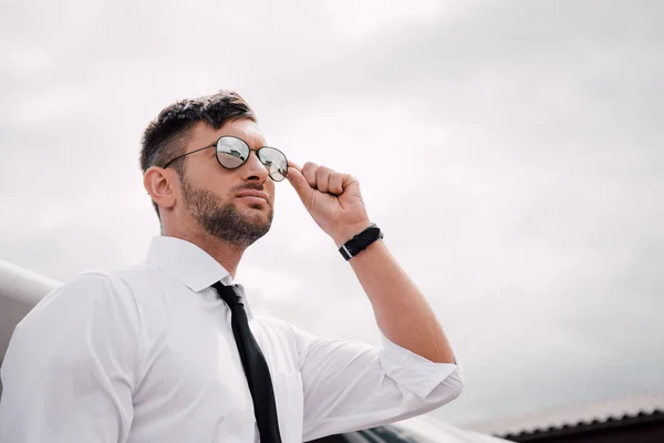 Vista de ángulo bajo del hombre barbudo confiado en desgaste formal y gafas de sol mirando hacia otro lado bajo el cielo nublado - foto de stock