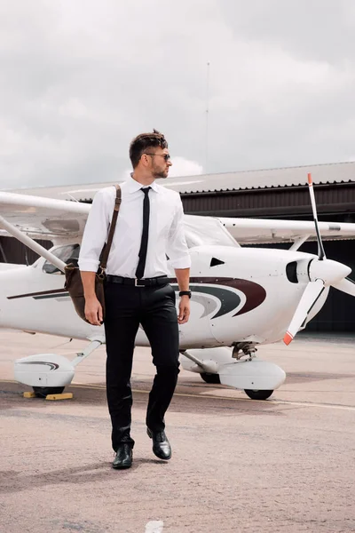 Full length view of confident pilot with bag standing near plane under cloudy sky — Stock Photo