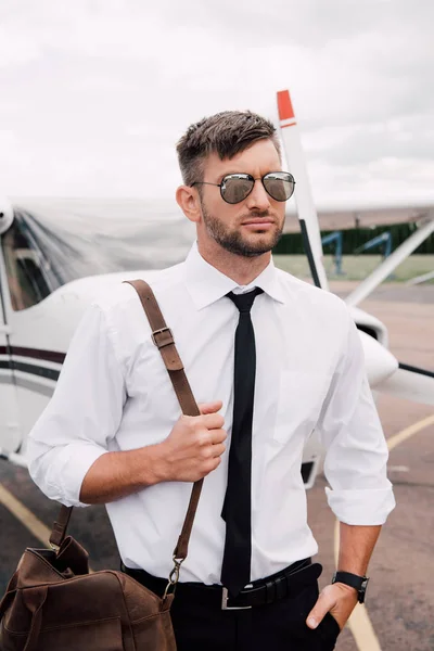 Confident pilot in sunglasses with bag standing with hand in pocket near plane — Stock Photo