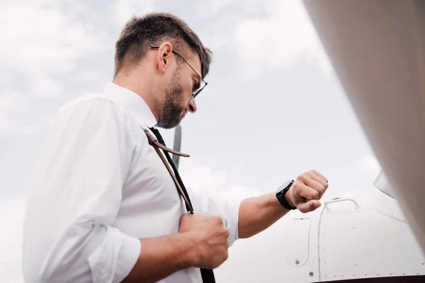 Vue à angle bas du pilote barbu dans des lunettes de soleil debout près de l'avion et regardant la montre-bracelet — Photo de stock