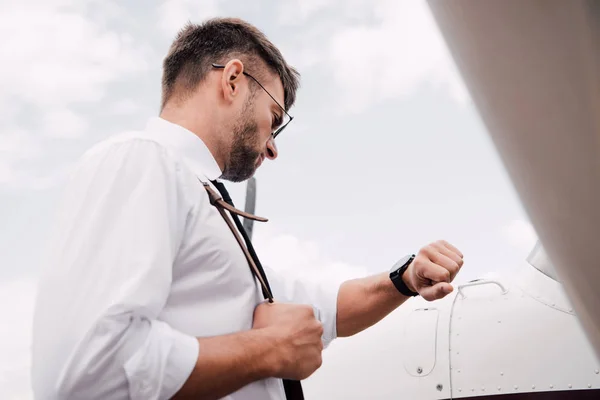 Vista de ángulo bajo del piloto barbudo en gafas de sol de pie cerca del avión y mirando el reloj de pulsera — Stock Photo