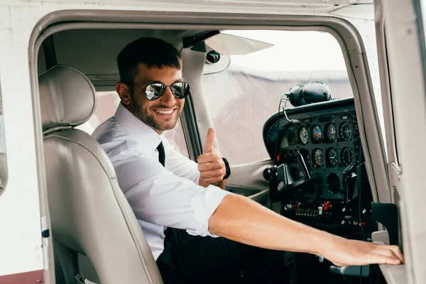 Улыбающийся красивый пилот в солнечных очках смотрит в камеру и показывает большой палец вверх в самолете — стоковое фото