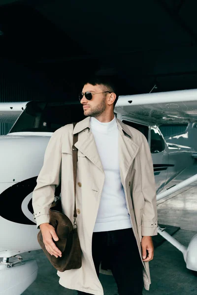 Pensativo hombre de negocios con estilo en abrigo de pie cerca de avión y mirando hacia otro lado - foto de stock
