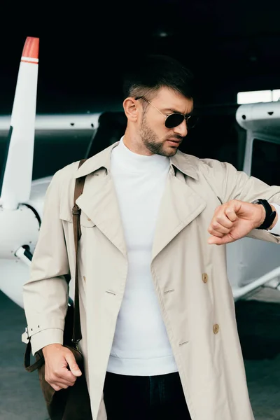 Elegante hombre de negocios en abrigo de pie cerca de avión y mirando reloj de pulsera - foto de stock