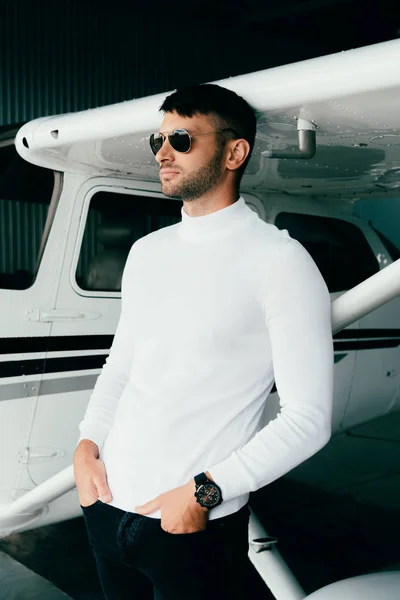 Schöner junger Mann in lässigem Outfit, die Hände in den Taschen in der Nähe des Flugzeugs und wegschauend — Stockfoto