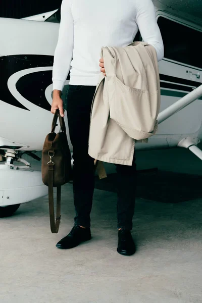 Vue partielle du jeune homme tenant manteau et sac alors qu'il se tenait près de l'avion — Photo de stock