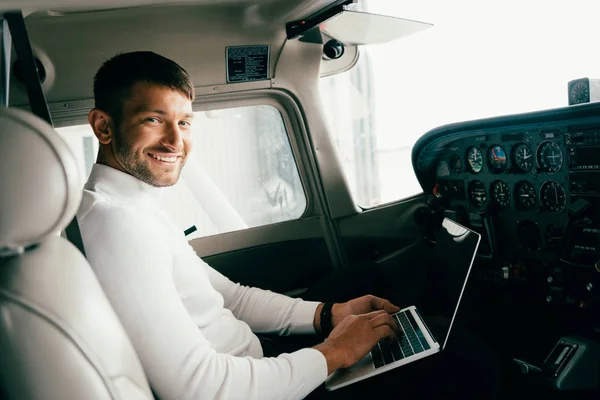 Smiling bearded young man in casual attire using laptop in plane — Stock Photo