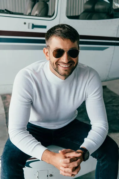 Smiling man in sunglasses sitting with clenched hands near plane — Stock Photo