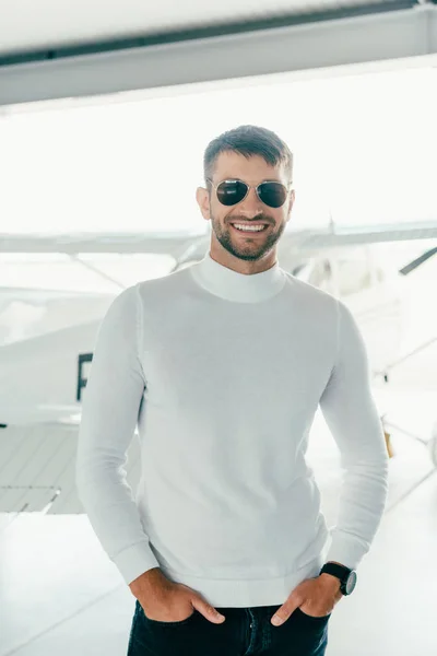 Vista frontale dell'uomo barbuto sorridente in occhiali da sole in piedi con le mani in tasche vicino all'aereo — Foto stock