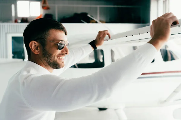 Vista lateral do homem barbudo sorridente em óculos de sol de pé perto do avião — Fotografia de Stock