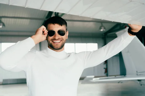 Vorderansicht eines lächelnden bärtigen Mannes mit Sonnenbrille, der in der Nähe eines Flugzeugs steht — Stockfoto
