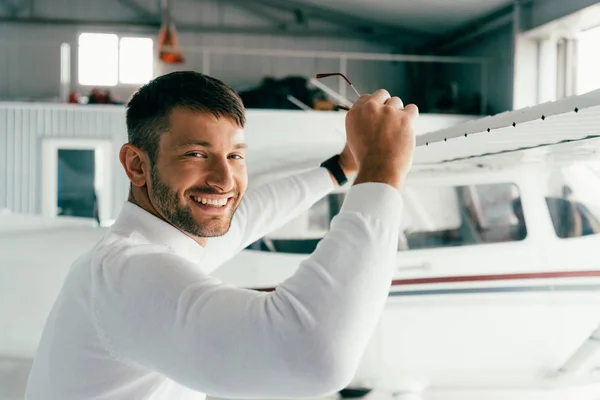 Uomo barbuto sorridente in piedi vicino all'aereo e tenendo gli occhiali da sole — Foto stock