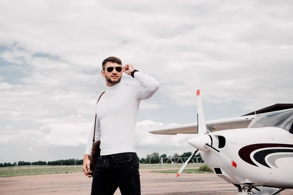 Uomo in occhiali da sole con borsa in piedi vicino aereo sotto cielo nuvoloso — Foto stock