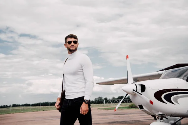 Homem em óculos de sol com saco de pé perto do avião sob céu nublado — Fotografia de Stock