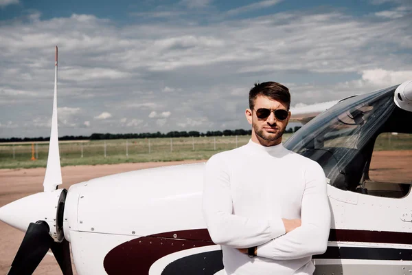Front view of man in sunglasses standing with crossed arms near plane in sunny day — Stock Photo