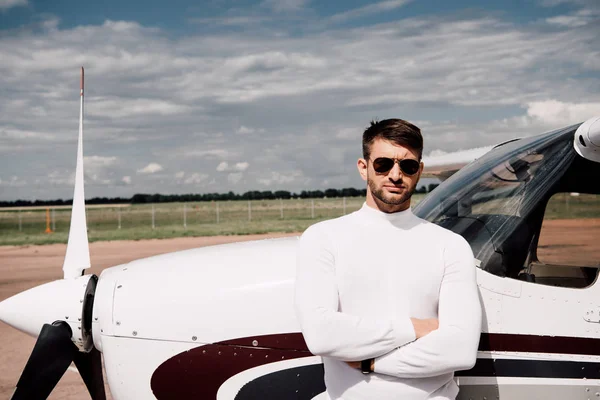 Frontansicht eines Mannes mit Sonnenbrille, der bei sonnigem Tag mit verschränkten Armen in der Nähe des Flugzeugs steht — Stockfoto