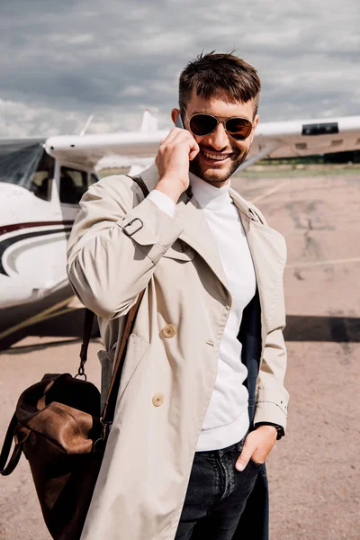 Улыбающийся мужчина в пальто с сумкой разговаривает на смартфоне рядом с самолетом — стоковое фото