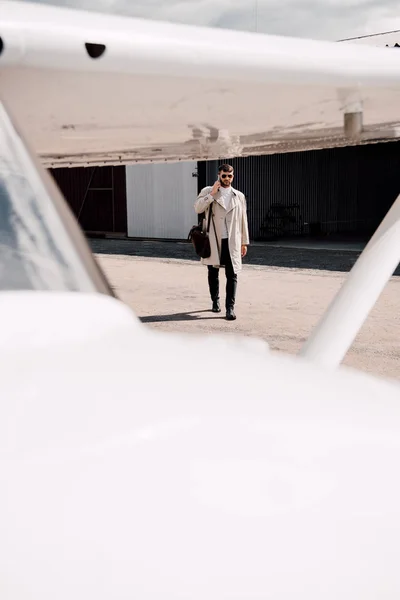 Ganzkörperansicht eines Mannes im Mantel mit Tasche, der bei sonnigem Wetter auf dem Smartphone in der Nähe des Flugzeugs spricht — Stockfoto