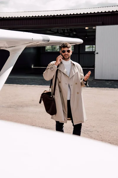 Uomo in cappotto con borsa che parla su smartphone vicino all'aereo nella giornata di sole — Foto stock