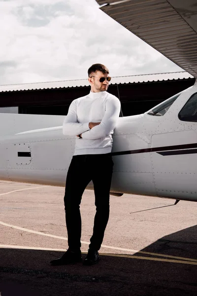 Vista completa del hombre serio en gafas de sol de pie con brazos cruzados cerca del avión - foto de stock