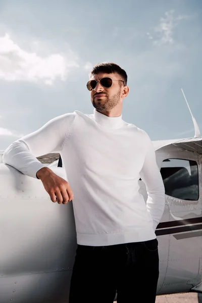 Vue à angle bas de l'homme barbu dans des lunettes de soleil debout près de l'avion dans la journée ensoleillée — Photo de stock