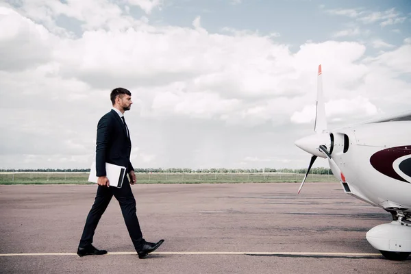 Vista completa de hombre de negocios en el desgaste formal celebración de la computadora portátil y carpeta cerca del avión en el día soleado - foto de stock