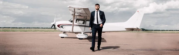 Panoramic shot of businessman in formal wear walking near plane in sunny day — Stock Photo
