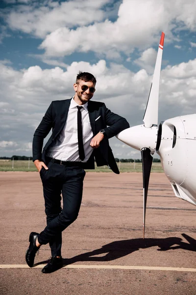Vue pleine longueur de souriant homme d'affaires en tenue formelle debout avec la main dans la poche près de l'avion — Photo de stock