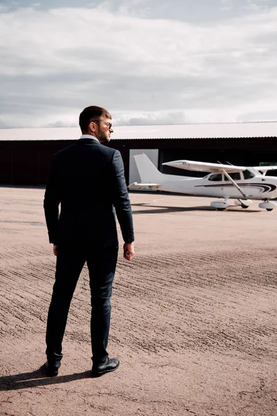 Vista trasera del hombre de negocios serio en ropa formal y gafas de sol de pie cerca de avión - foto de stock