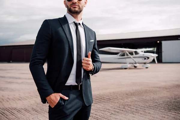 Vista parcial del hombre de negocios en ropa formal de pie con la mano en el bolsillo delante del avión y mostrando el pulgar hacia arriba - foto de stock