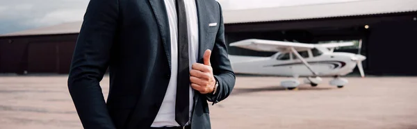 Panoramic shot of businessman in formal wear standing in front of plane and showing thumb up — Stock Photo