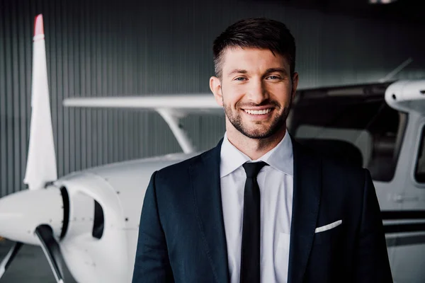 Vue de face de l'homme d'affaires souriant en tenue formelle debout près de l'avion et regardant la caméra — Photo de stock