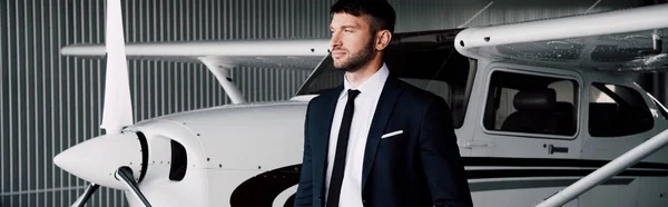 Panoramic shot of confident businessman in formal wear standing near plane and looking away — Stock Photo