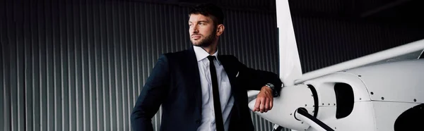 Panoramic shot of confident businessman in formal wear standing near plane and looking away — Stock Photo