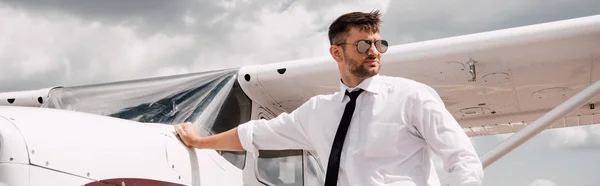 Panoramic shot of confident pilot in sunglasses standing near plane — Stock Photo