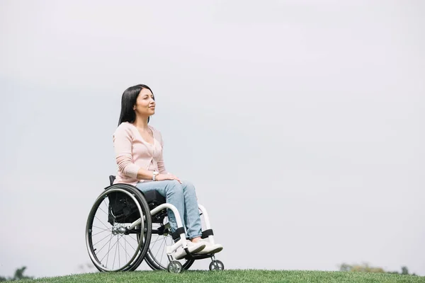 Mujer discapacitada feliz mirando hacia otro lado mientras está sentada en silla de ruedas en el parque - foto de stock
